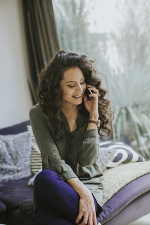 Curly hair young woman using mobile phone and holding mug while sitting on sofa in the roomの写真素材