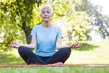 Senior woman in lotus pose sitting on green grass at summar dayの写真素材
