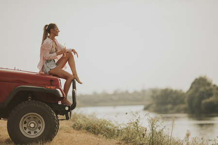 Young woman sits on a car hood,  drinks refreshment and enjoys itの写真素材