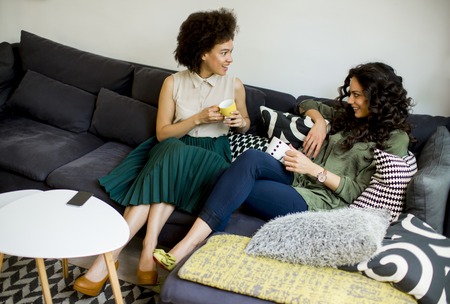 Two multiracial young women chatting and drinking coffe in rhe living roomの写真素材