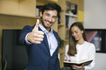 Portrait of young businessman with thumb up standing with businesswoman in officeの写真素材