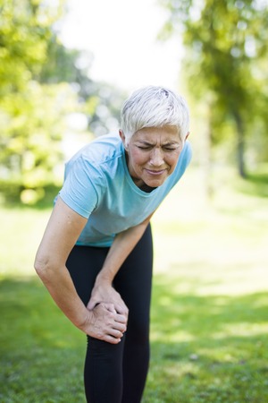 Senior sportswoman with knee pain standing in park on summer dayの写真素材