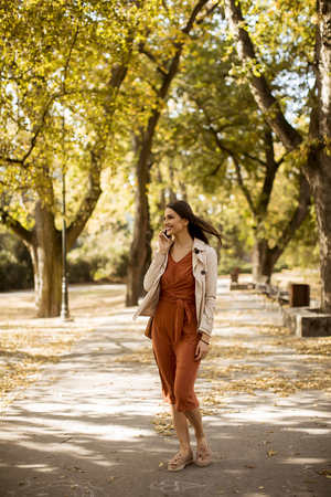 Happy young woman using cell phone in autumn park on beautiful dayの写真素材