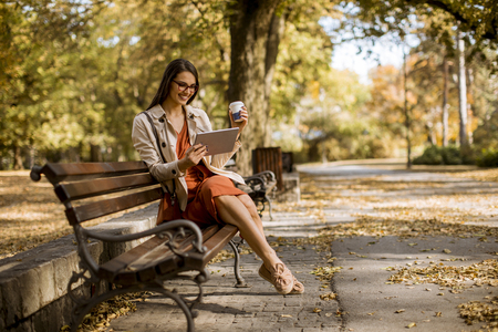 Woman drinks coffee and  sitting on bench in park during autumn weather, using tablet pc and  checking social media.の写真素材