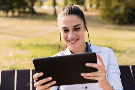 Young woman sitting on the bench in the park and listen music on  a tabletの写真素材
