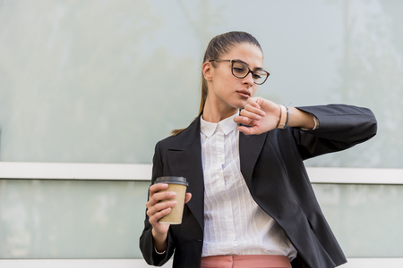 Portrait of businesswoman in front of building looking on wristwatch and drinks coffeeの写真素材