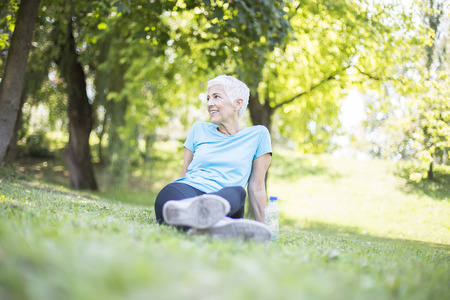 Senior woman sitting and resting after workout in park on grassの写真素材
