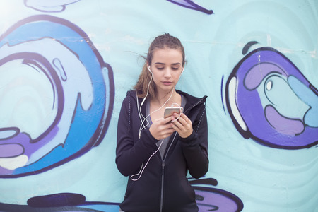 Portrait of female jogger taking break and  listening music on mobile phone in front of graffiti...