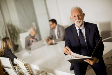 Portrait of senior businessman standing in office while other business people having meetingの写真素材