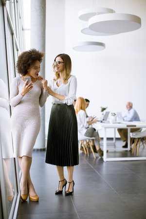Two young businesswomen with a mobile phone in the office and other business people working at a deskの写真素材