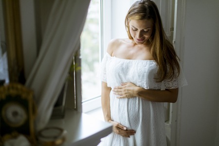 Pretty young pregnant woman standing by the windowの写真素材