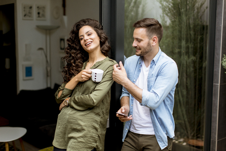 Happy young cute couple in love embracing each other and drinking coffee at home by the windowの写真素材