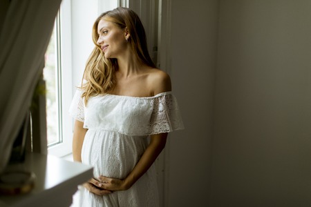 Pretty young pregnant woman standing by the windowの写真素材