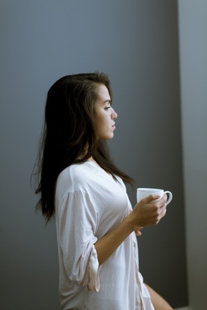 Portrait of attractive young woman with mug by the window in bedroom