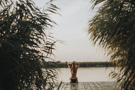 Back view at attractive young woman in bikini sitting on a pier at the lakeの写真素材