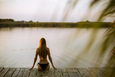 Back view at attractive young woman in bikini sitting on a pier at the lakeの写真素材