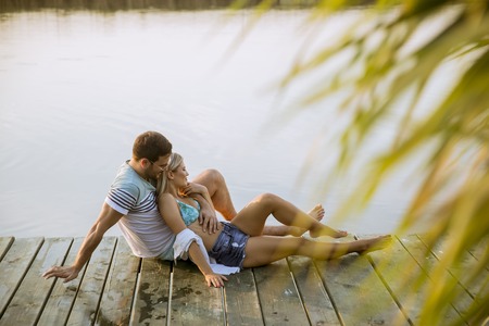 Loving couple sitting on the pier on lake at summer sunsetの写真素材