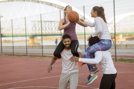 Young women sitting on the men shoulders and holding a basketball at outdoor court on a sunny dayの写真素材