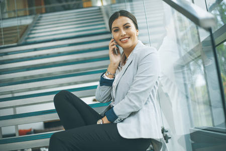 Pretty young business woman sitting on the stairs at the office and use mobile phoneの写真素材