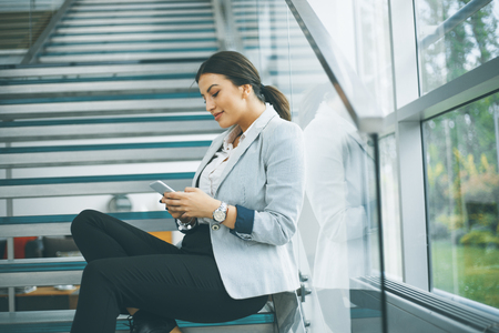 Pretty young business woman sitting on the stairs at the office and use mobile phoneの写真素材