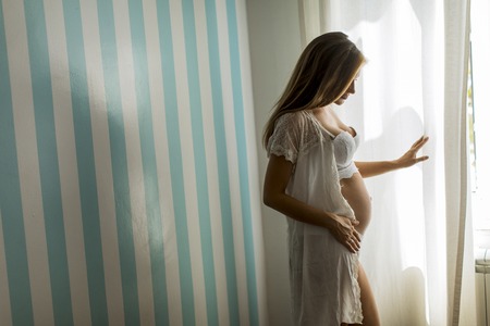 Pretty young pregnant woman standing at window by the blue stripped wall in the room at daylightの写真素材