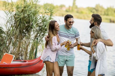 Group of friends with cider bottles standing by the boat near the beautiful lake and having fun on a summer dayの写真素材