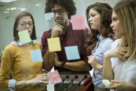 Group of young business people discussing in front of glass wall using post it notes and stickers at...
