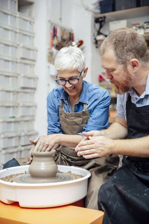 Senior woman spinning clay on a wheel with a help of a teacher at pottery class