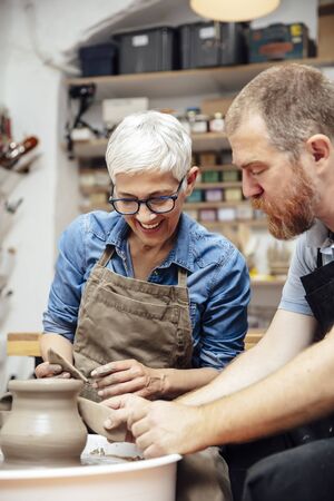 Senior woman spinning clay on a wheel with a help of a teacher at pottery classの写真素材