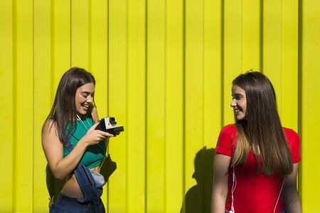 Two pretty young happy women taking photo with retro camera and have a great time by yellow wallの写真素材