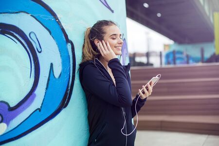 Young female jogger listening music on mobile phone in front of graffiti wall