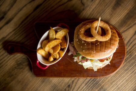 Beef burger in homemade pizza dough bun with BBQ sauce, melted chedar cheese, bacon, tomato, rocket, lettuce with onion rings and spicy fries on the tableの写真素材