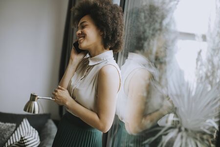 Pretty young woman with curly hair using mobile phone by the window at homeの写真素材