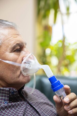Senior man using medical equipment for inhalation with respiratory mask, nebulizer in the room