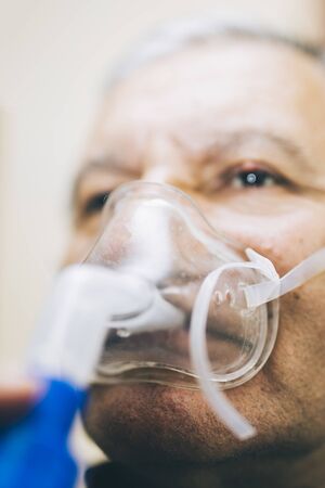 Senior man using medical equipment for inhalation with respiratory mask, nebulizer in the room