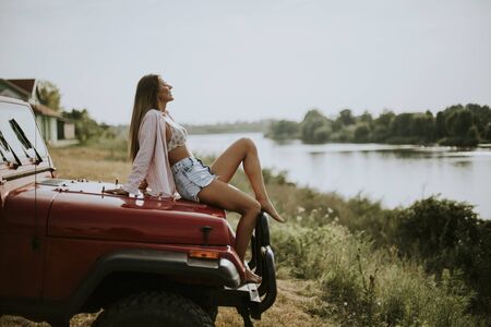 Pretty young woman sits on a red car hood and enjoys in summer sunの写真素材