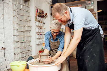 Senior woman spinning clay on a wheel with a help of a teacher at pottery class