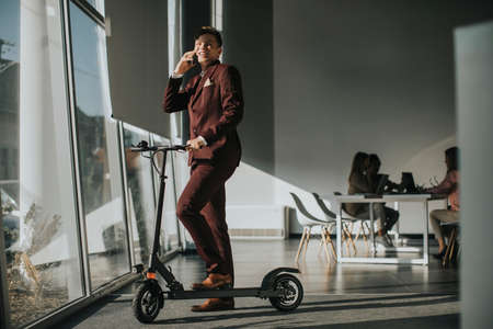 Handsome young businessman standing by the office window with electric scooter and using mobile...