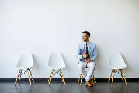 Young man sitting at chair in the waiting room with a folder in hand and checking time before an interviewの写真素材