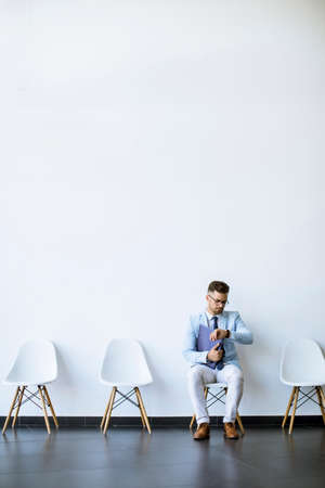 Young man sitting at chair in the waiting room with a folder in hand and checking time before an interviewの写真素材