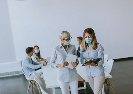Two businesswomen, mature and young one, talking in the office and wearing mask as a virus protectionの写真素材