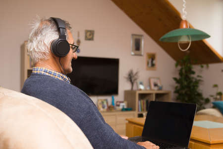 Modern elderly man siting at home with earphones and having online video call