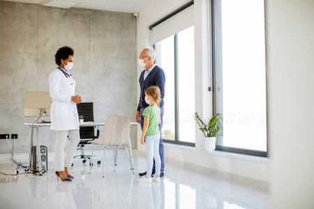 Grandfather with protective facial masks with cute granddaughter at black female doctorの写真素材