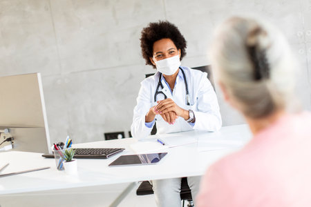 Senior woman with protective facial masks receive news from black female doctor in the officeの写真素材