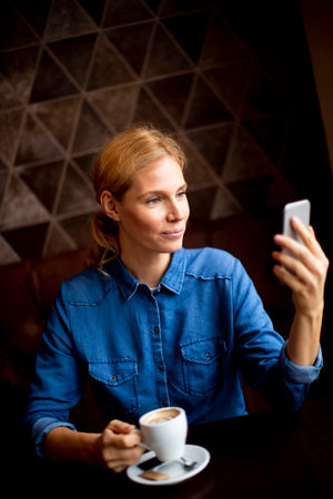 Pretty young woman sitting at a table with coffee or cappuccino and using a mobile phone