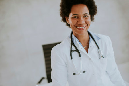 Female African American doctor wearing white coat with stethoscope standing by desk in the office