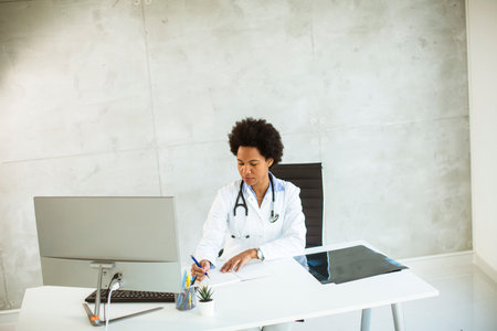 Female African American doctor wearing white coat with stethoscope sitting behind desk in the office