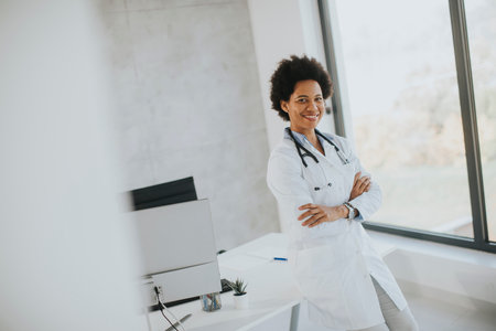 Female African American doctor wearing white coat with stethoscope standing by desk in the officeの写真素材
