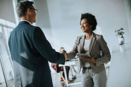 African American businesswoman holding digital tablet and looking at handsome colleague while shaking hands in officeの写真素材