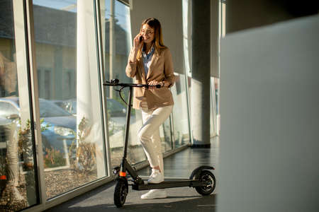 Pretty young woman standing by the office window with electric scooter and using mobile phoneの写真素材